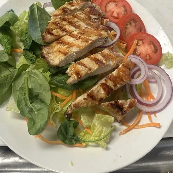 A grilled chicken salad plated with spinach, romaine, sliced tomato, red onion and shredded carrot.