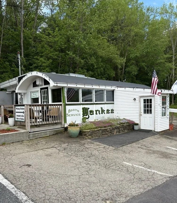 The front of Yankee Diner: green Old-English 'Yankee' lettering and a 'Booth Service' sign on white clapboard, with flowers along a low stone wall.