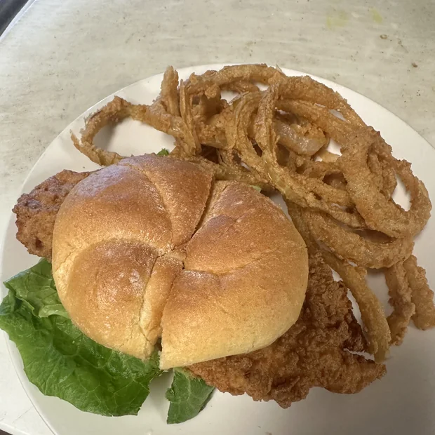 A crispy chicken breast sandwich on a bulkie roll with lettuce, piled alongside a nest of homemade onion rings.