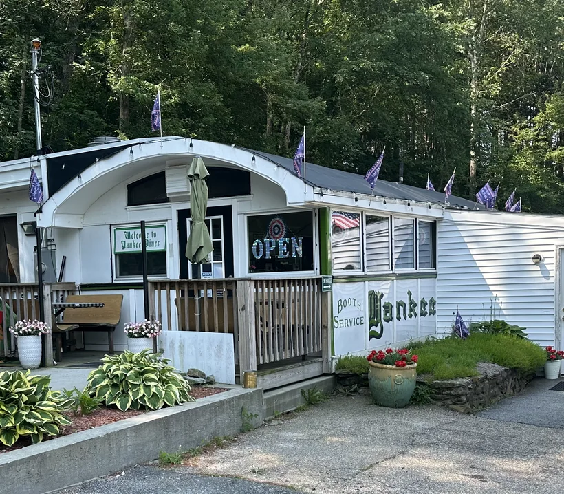 The diner entrance: a small porch with an OPEN sign, hostas and potted flowers along a low stone wall, and Yankee painted in green on the white clapboard.