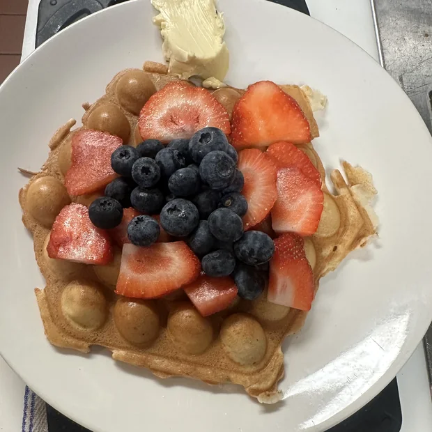 A waffle dusted with powdered sugar and topped with sliced strawberries, blueberries and a pat of butter.